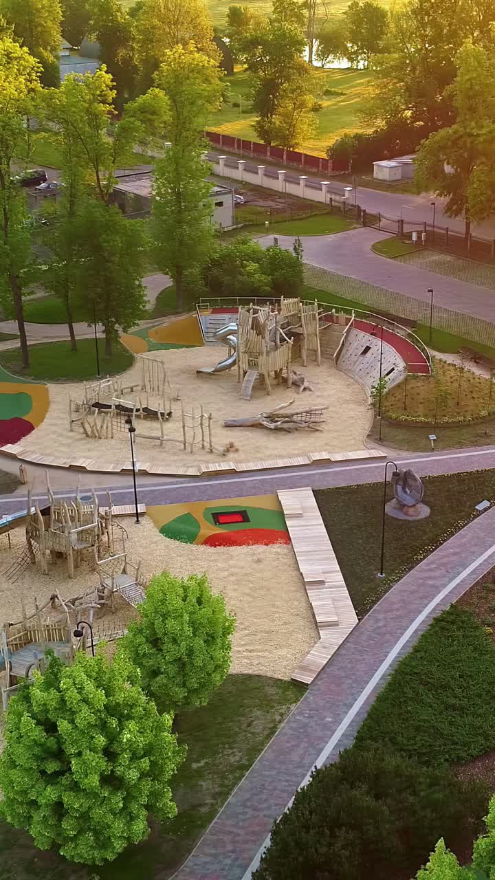 A peaceful aerial flyover shows an empty kids playground in Latvia at sunset, featuring modern climbing equipment, slides, sand pits and green park surroundings bathed in golden light - vertical