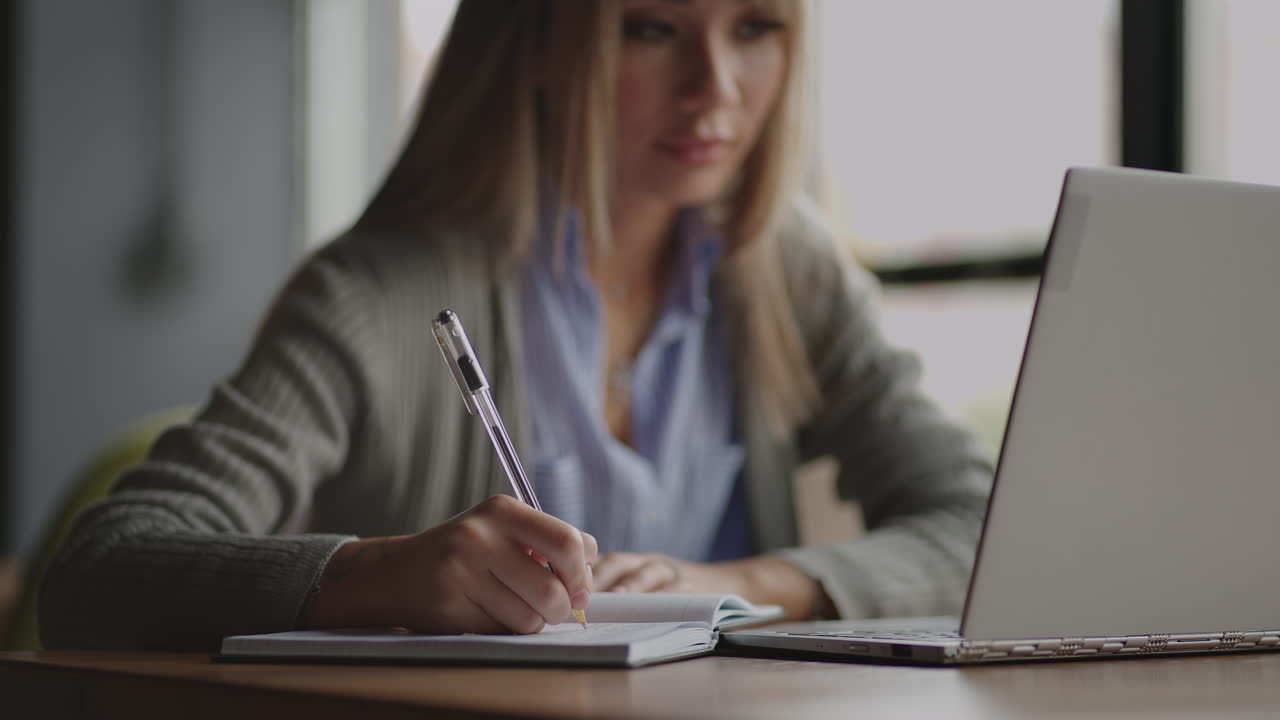 Asian woman working on her laptop and writing in his notebook sitting at a table. Working in coffee shop. woman looking to a laptop screen and making notes in her notebook. studying online.