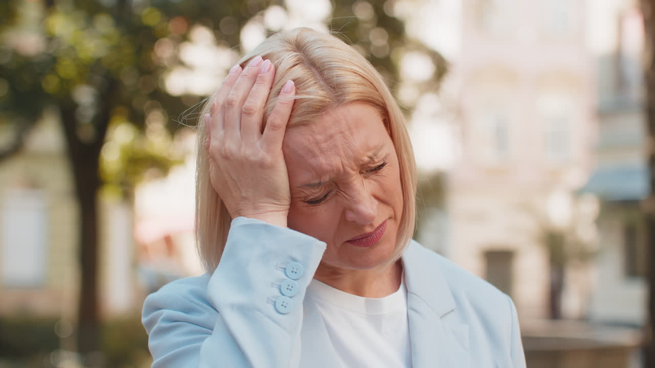 Sick mature caucasian businesswoman in formal suit feels headache while standing on city street