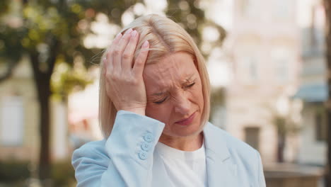 sick mature caucasian businesswoman in formal suit feels headache while standing on city street