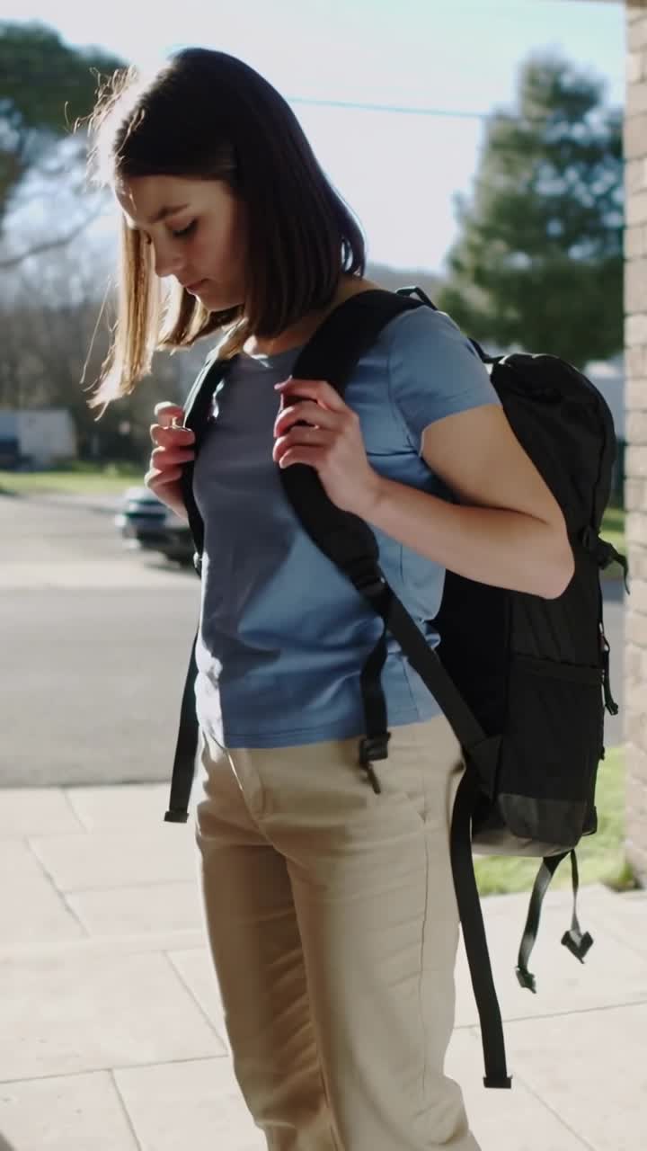 Vertical video: Adjusting woman in blue shirt securing black backpack straps by pillar, ready to go