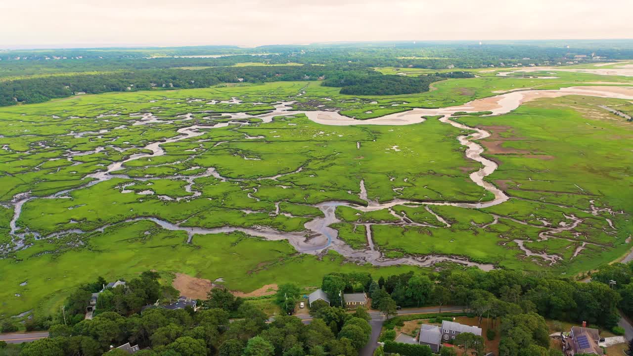 Aerial view of expansive wetlands shows winding tidal creeks and reflective pools, lush green grasses spread across the marsh, and sandy edges blending into the coastal horizon