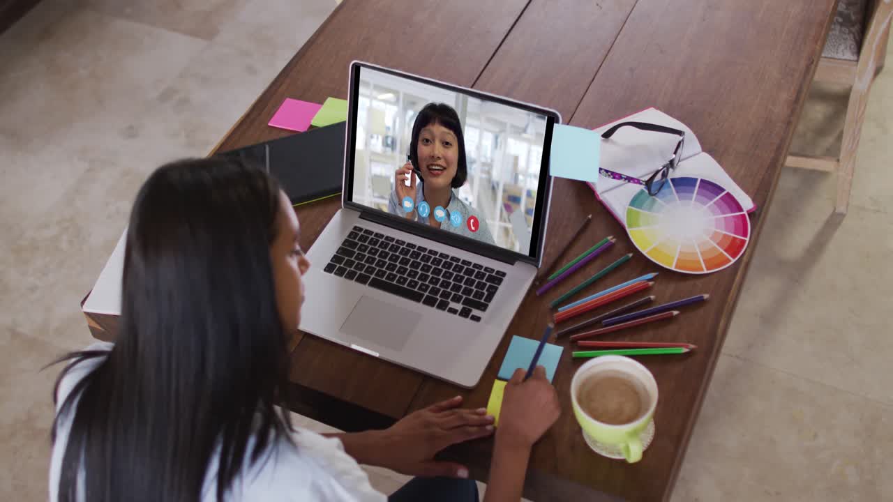 Mujer caucásica usando una laptop en videollamada con una colega y tomando notas