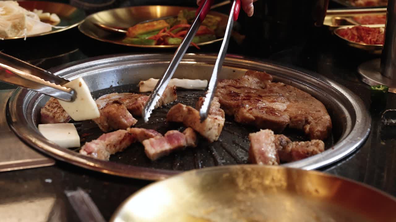 Hands grill pork belly slices with tongs on tabletop Korean BBQ under warm ambient lighting