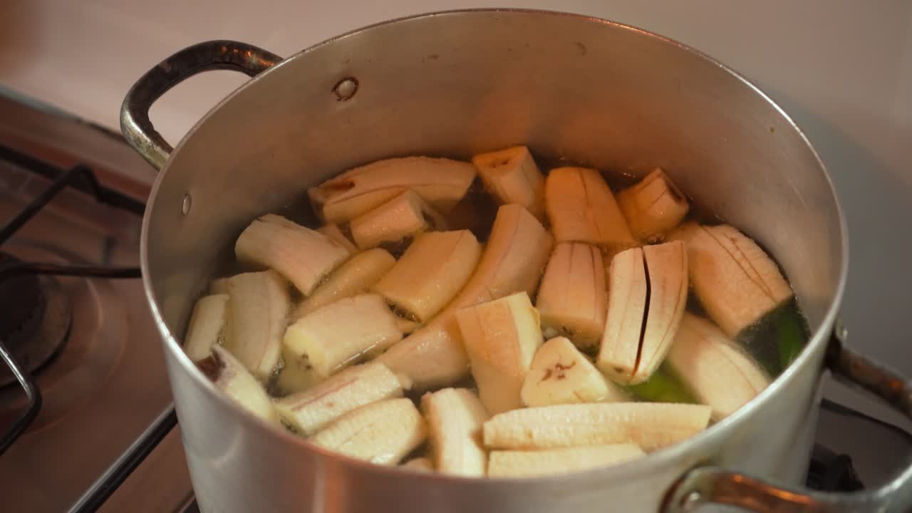 Chunks of green plantain cooking in boiling water inside a large silver pot on stovetop. Latin American cuisine. Ideal for food content, recipes, or kitchen footage.