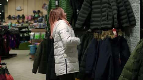 Woman-is-fitting-a-new-down-jacket-from-a-rack,-looks-at-the-mirror