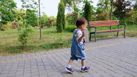 following the cute running toddler in the park. baby in a jeans romper runs holding hands up.