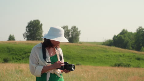 female photographer wearing white hat and green dress walking in tall grassy meadow under bright sky while holding camera, ready to take photo in calm rural setting filled