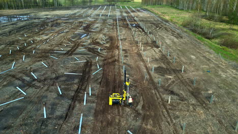 Aerial-view-tilitng-toward-a-worker-installing-poles-at-a-solar-park,-spring-day