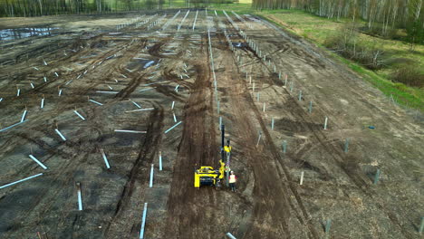 aerial view tilitng toward a worker installing poles at a solar park, spring day