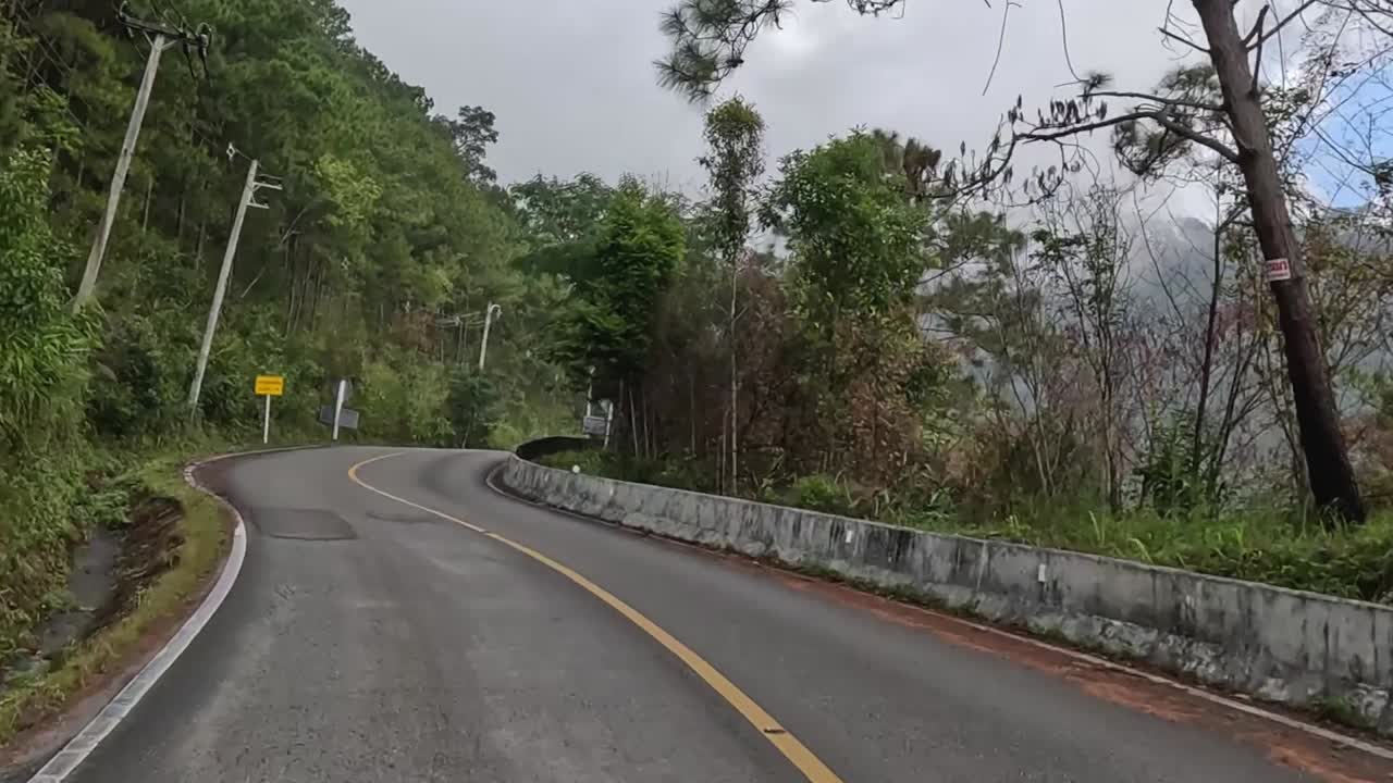 A winding road surrounded by dense greenery and misty mountain views, featuring caution signs and road markings.