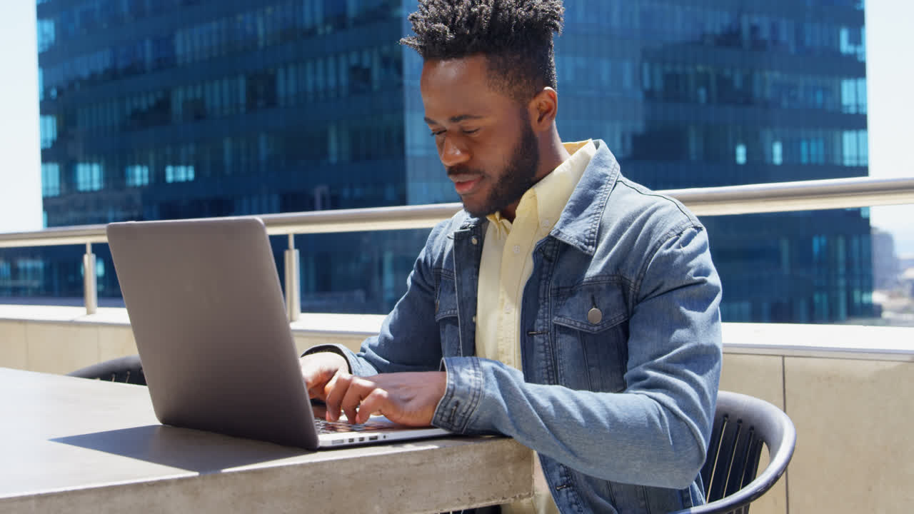 Side view of young cool black businessman working on laptop in balcony of modern office 4k