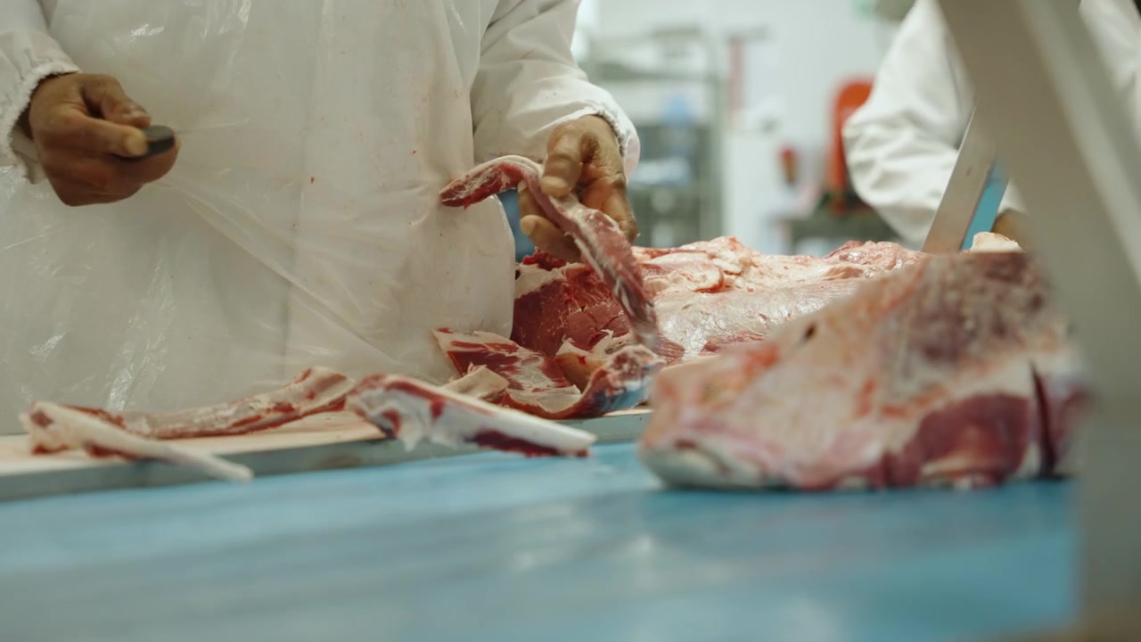Gloved hands trim bone and excess fat from a raw carcass joint on a blue cutting bench in an industrial butchery.