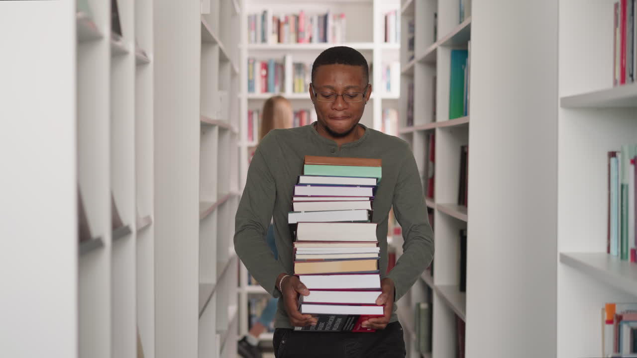 Black man with book stack in library. African American customer carries large literature pile walking along aisle between bookcases. Information learning