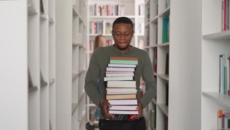 hombre negro con una pila de libros en la biblioteca. cliente afroamericano lleva una gran pila de literatura caminando por el pasillo entre las librerías. aprendizaje de información