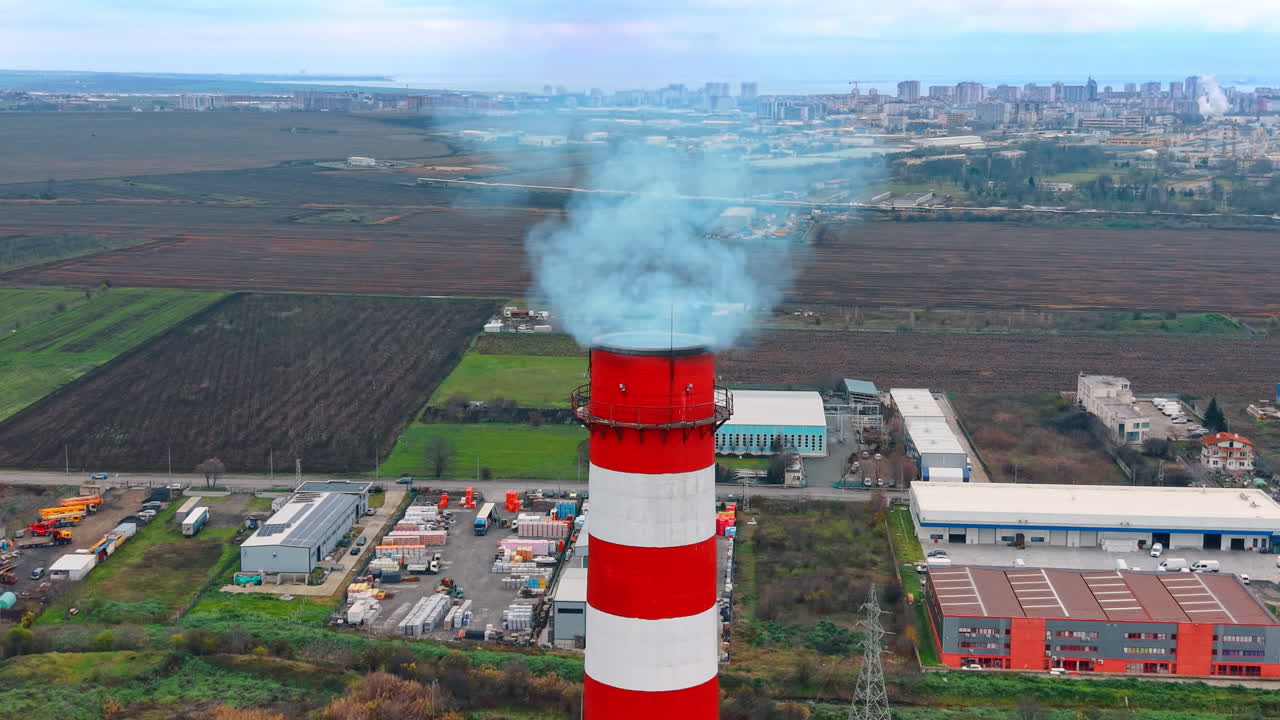 Ciminiera industriale che emette fumo con città e mare sullo sfondo. Camino a strisce rosse e bianche che rilascia scarico blu su un quartiere di magazzini e un'area urbana