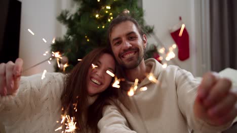 Portrait-of-a-happy-couple,-a-guy-and-a-girl-in-white-sweaters-hold-sparklers-in-their-hands,-rejoice-at-the-upcoming-Christmas-holiday-and-have-fun-in-the-cozy-atmosphere-of-a-house-decorated-in-Christmas-style