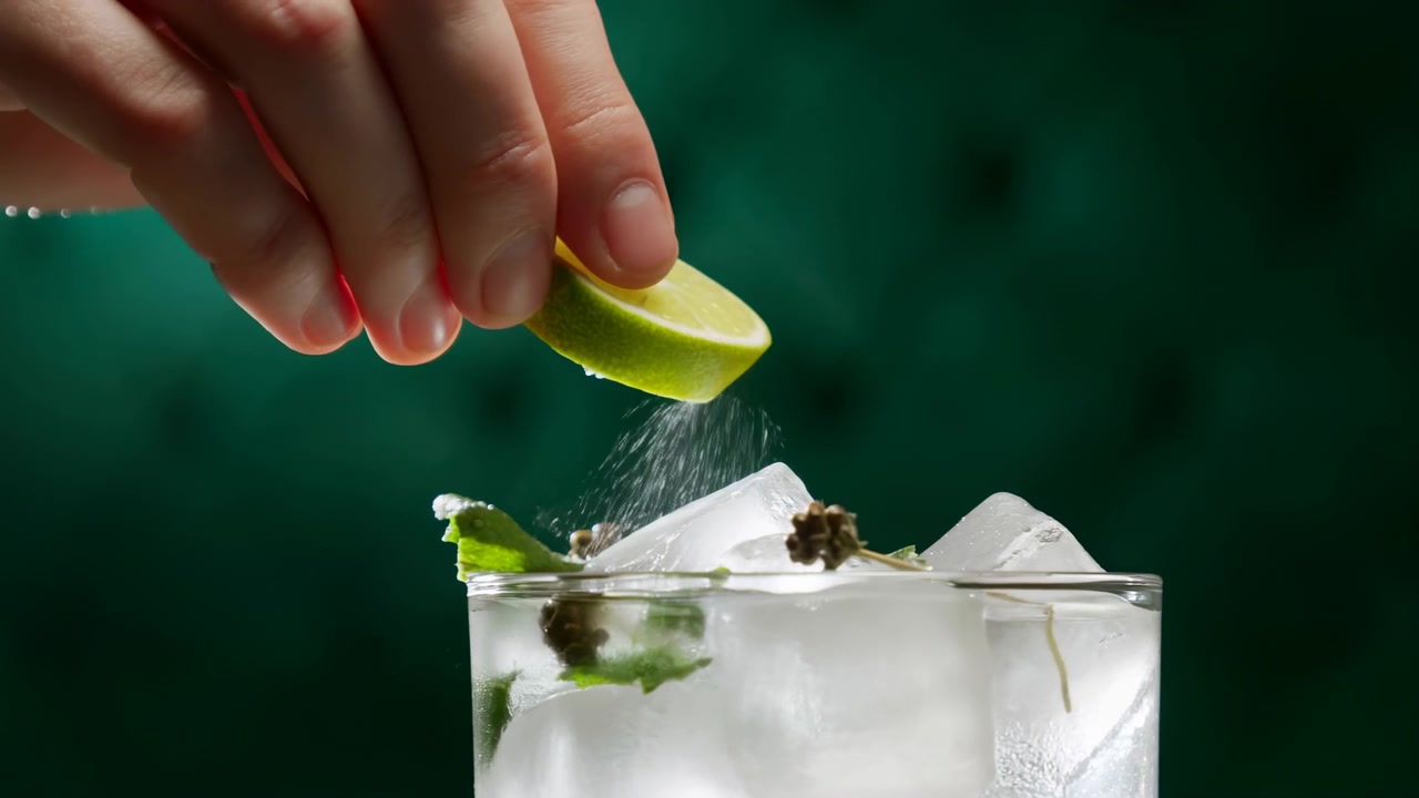 Hand holds lime wedge above clear iced cocktail, releasing fine citrus dust over mint leaves, herb garnish, and bright ice cubes in short glass with moody green backdrop and fresh styling