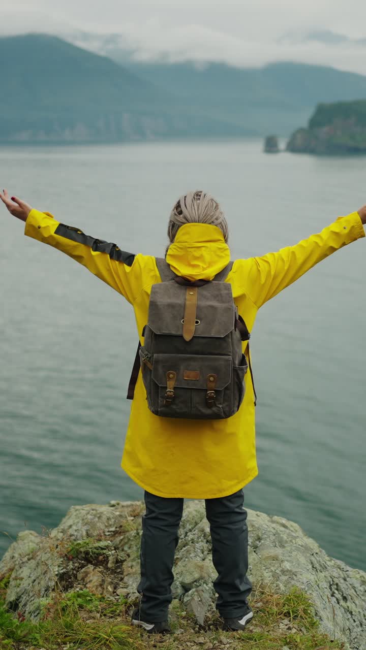 Donna con impermeabile giallo e zaino in piedi sul bordo di una scogliera che si affaccia sul mare