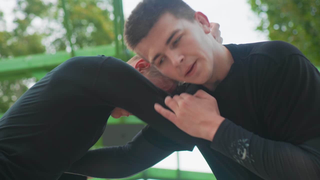 close clinch between two white men focused control drill emphasizing neck hold escape technique coachlike instruction leafy outdoor gym backdrop concentrated athletic intensity restrained pace
