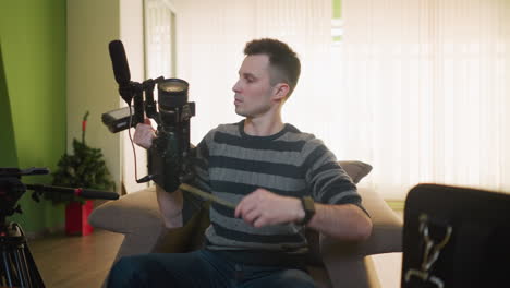 studio guy holding camera with microphone attached, focusing on lens. preparing for video shoot in home studio. camera gear, microphone, and other accessories set up on table