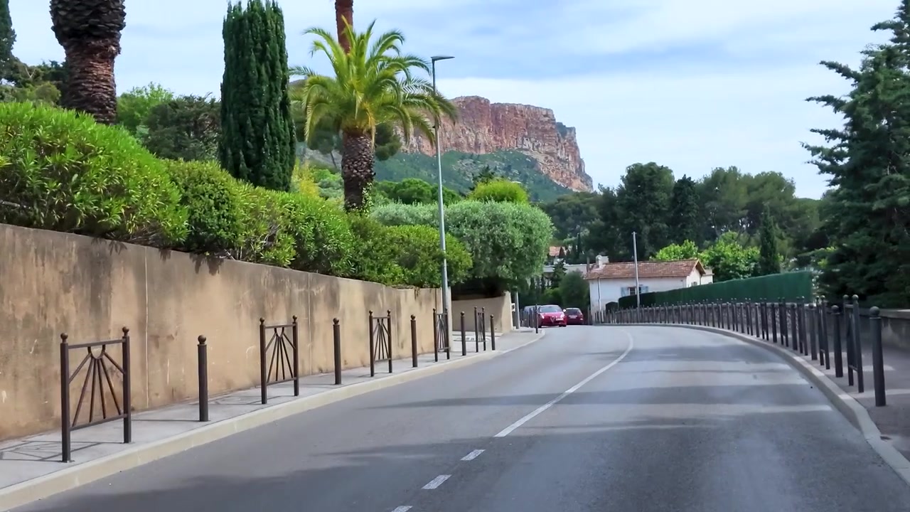 Una telecamera panoramica lungo una strada asfaltata curva, oltre muri in pietra e vegetazione mediterranea