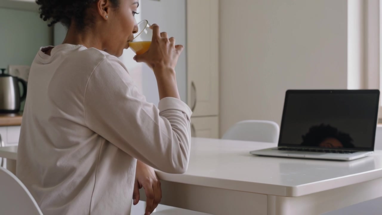 Donna con top color cuoio che allunga la mano afferrando un bicchiere di succo d'arancia, sorseggiando e controllando il laptop al tavolo della cucina
