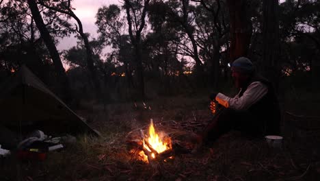 a bushman sits around a campfire in the sunset glow out in the australian bush
