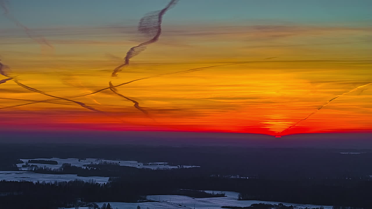 Eine seitliche Luft-Hyperlapse-Aufnahme fängt die glühende rote und goldene Sonne ein, die unter dem gefrorenen lettischen Horizont versinkt, während sich dramatische Flugzeugkondensstreifen über einen lebhaften, feurigen Winterhimmel schlängeln.