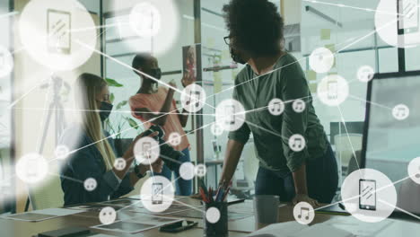 Animation-of-connected-icons-over-diverse-female-coworkers-wearing-mask-discussing-reports-in-office