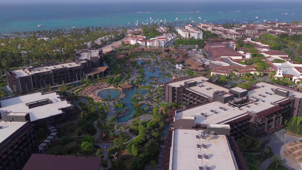 Ampia veduta aerea che curva a sinistra sopra un complesso alberghiero tropicale, con piscine, giardini lussureggianti, attrazioni acquatiche e il Mar dei Caraibi