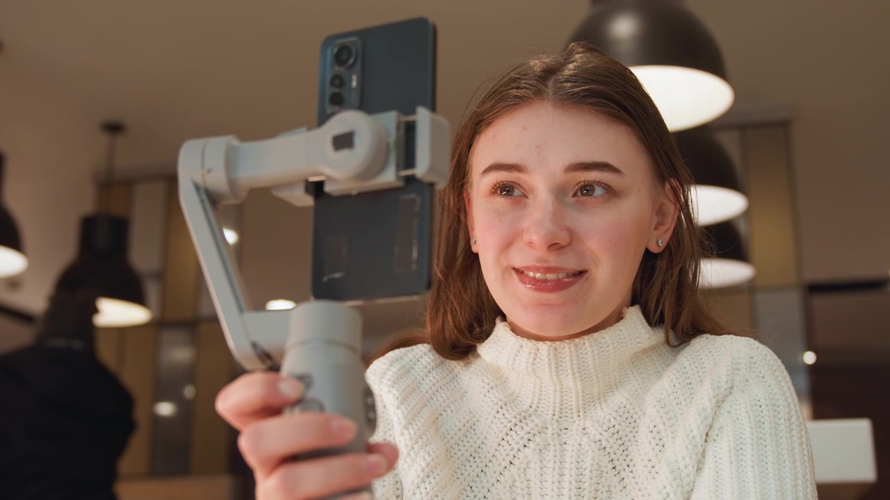 Ung kvinna använder smartphone-gimbal på ett mysigt kafé, justerar den handhållna stabilisatorn medan hon pratar, ler och skapar engagerande socialt innehåll i varmt naturligt ljus med en avslappnad inomhusatmosfär.