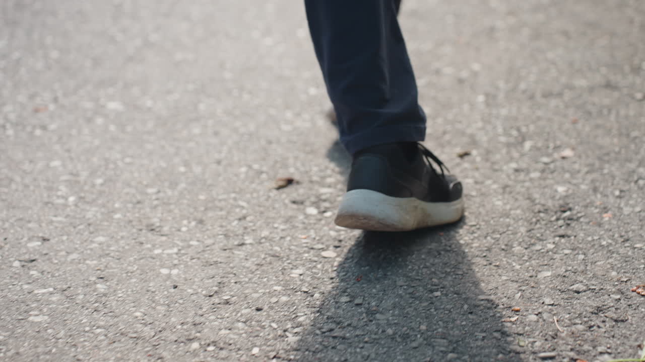 Güneş ışığıyla aydınlanan kaldırımda yakın çekim spor ayakkabılarla rahat bir şekilde yürüyüş, gölge pürüzlü asfalt üzerinde uzanıyor, kot pantolonlu bacaklar sabit adımlarla hareket ediyor, bağcıklar yerde sürtünüyor, rahat bir öğleden sonra şehirde gezinti