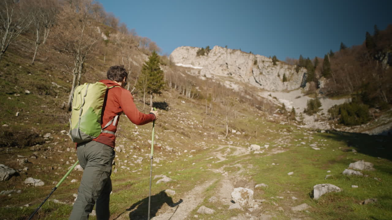 seguimiento de cámara desde detrás de un excursionista caminando por un sendero en una montaña, tees deciduoud sin hojas, cielo despejado