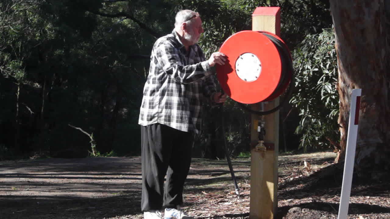A man approaches and operates a fire hose reel located in a green outdoor area. The scene showcases an essential safety feature amidst nature, highlighting preparedness and fire safety