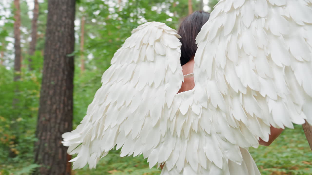 Actress dressed in white angel costume turns around in lush forest, adjusting hair while holding wooden staff, surrounded by soft green light and tall trees