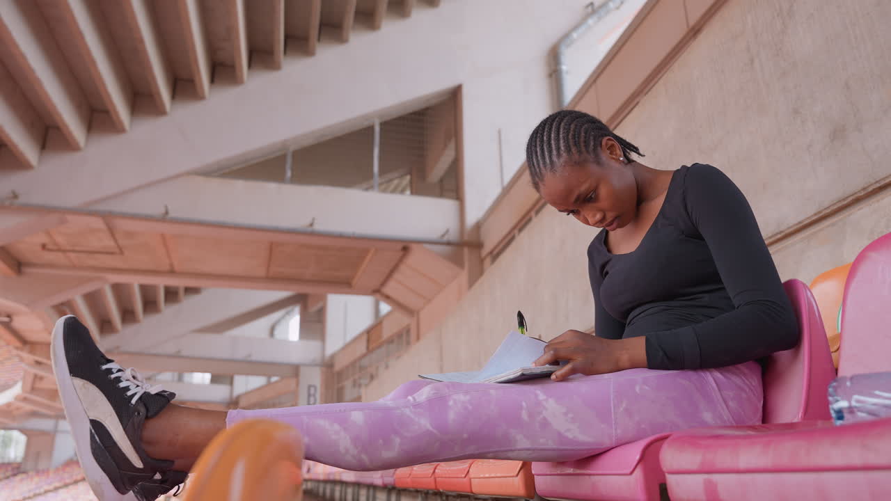 wide shot black woman on bleachers using phone, pink seats and stadium roof framing scene, purple pants and sneakers, casual streetwear mood, scrolling and waiting in empty urban venue