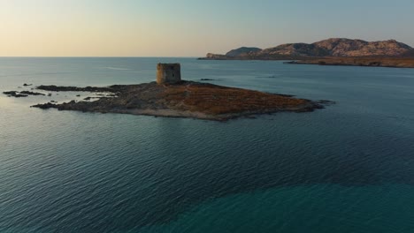 Aerial-of-La-Pelosa,-a-famous-natural-sand-beach-sandy-bay-on-the-tourist-holiday-island-Sardinia-in-Italy-by-sunset-with-clear-blue-turquoise-water-and-a-lighthouse-watchtower