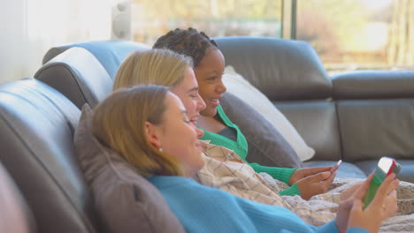 group of multi-cultural teenage girl friends snuggled under blanket looking at mobile phones at home