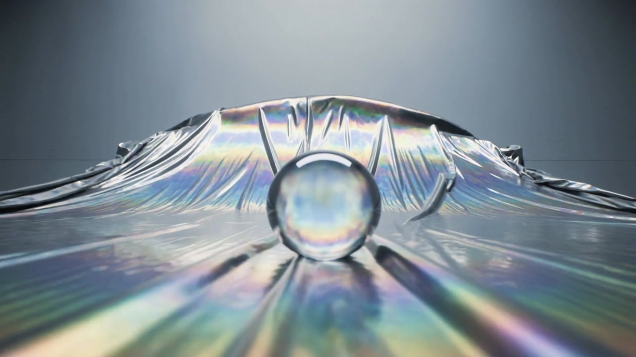 Glass orb pressing into iridescent sheet under spotlight in studio, forming dome with rainbow sheen