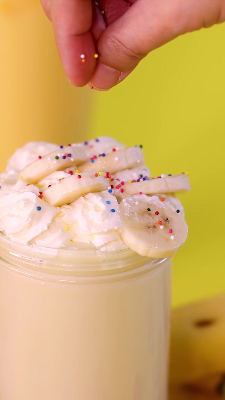 Mano che sparge granella colorata su milkshake alla banana con panna montata, sfondo giallo brillante, primo piano.