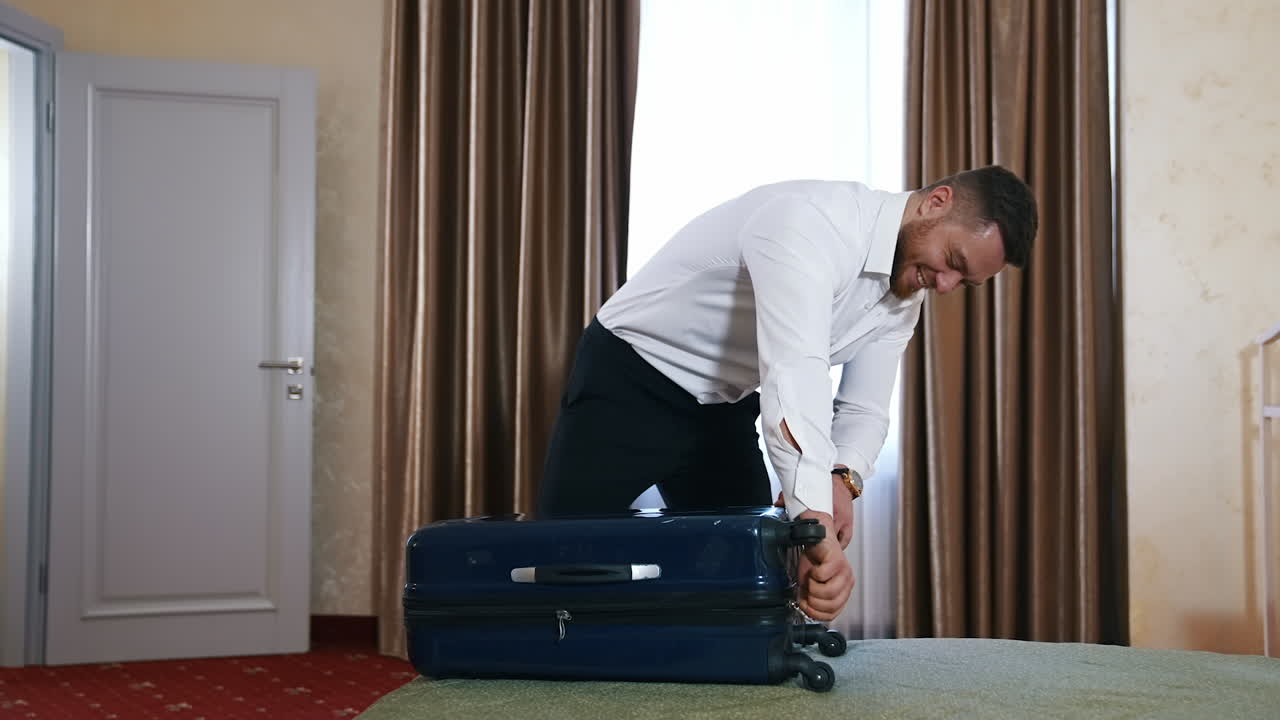 Happy businessman leaving hotel room. Young man in white shirt and trousers closes his luggage. Traveller finishes packing stuff and ready to leave hotel.