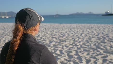 Vista-Trasera-De-Una-Mujer-Morena-Con-Gorra-Sentada-En-La-Arena-Y-Disfrutando-De-La-Vista-Al-Mar-En-La-Playa-De-Whitehaven-En-Queensland