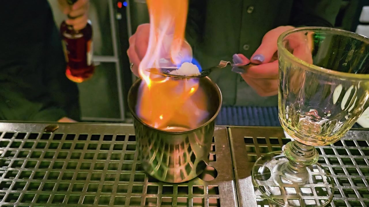 Cận cảnh một người pha chế đang chuẩn bị ly cocktail bốc lửa tại một quán bar thiếu sáng, với ngọn lửa xanh, viên đường, muỗng có rãnh, cốc kim loại, ly thủy tinh, quầy thép không gỉ và ánh sáng đêm huyền ảo.
