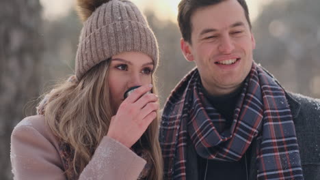 happy couple holding hot tea cups over winter landscape. young couple in love on a winter vacation, standing next to a tree and drinking hot cup of tea