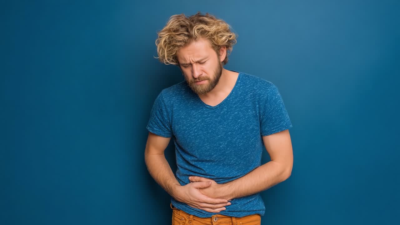 Uomo con i capelli ricci che prova disagio mentre si tiene lo stomaco su uno sfondo blu, mostrando un'espressione di dolore indicativa di un disturbo addominale