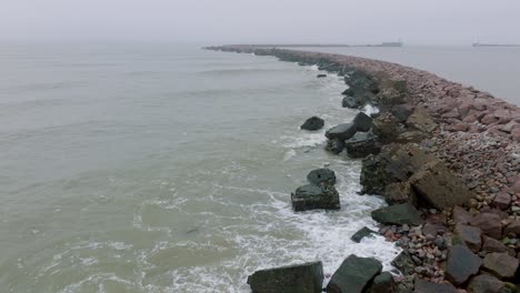 aerial establishing view of port of liepaja stone pier, baltic sea coastline , foggy day with dense mist, moody feeling, big storm waves splashing, wide drone shot moving forward low