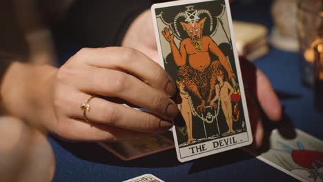 un primer plano de una mujer dando una lectura de tarot en una mesa a la luz de las velas sosteniendo la carta del diablo 2