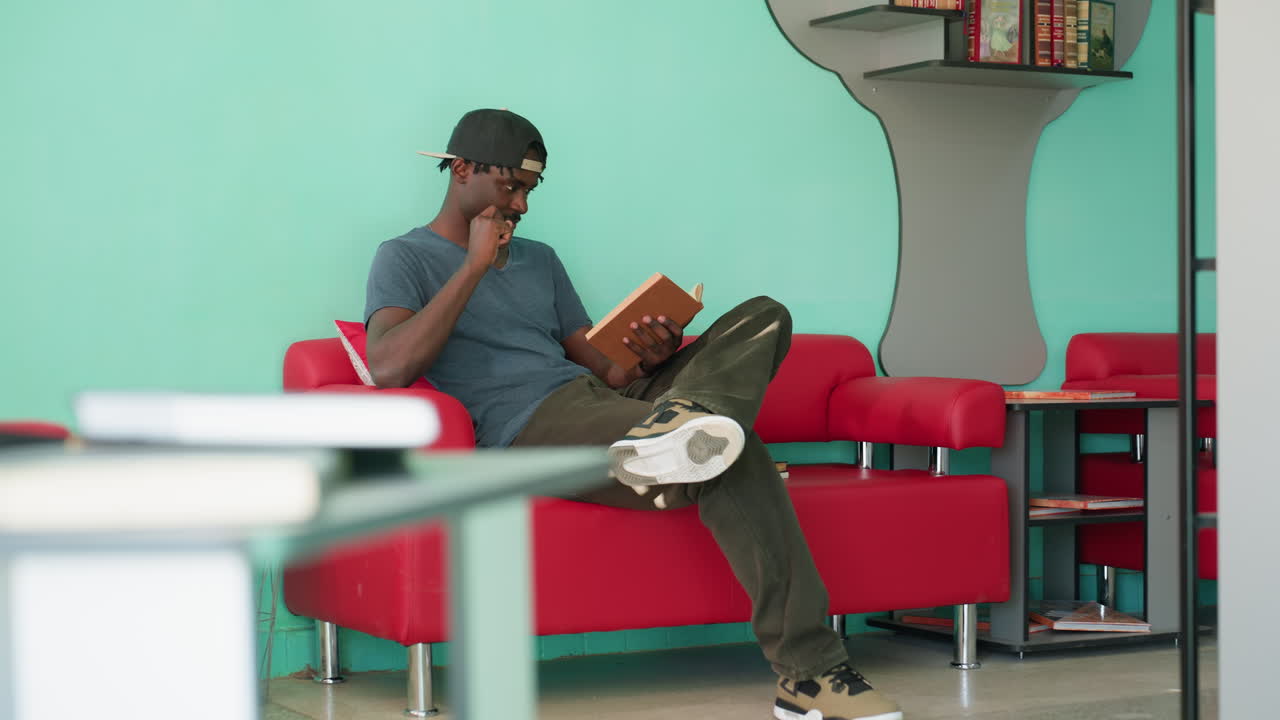 Relaxed adult in casual shirt and cap sits on red couch with crossed leg, holding open book while reading with focus in bright indoor space with turquoise wall and bookshelf