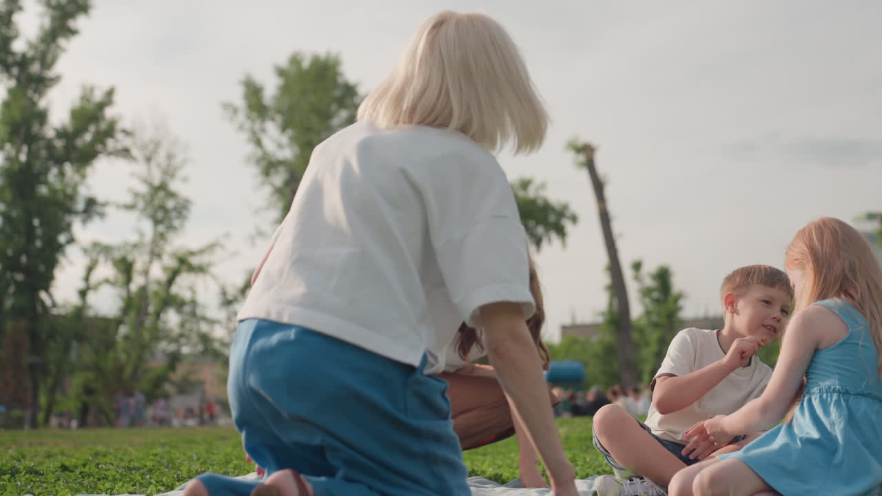 back view of young mothers with friend spreading blanket on green grass in sunny park with blurred background while children sit nearby creating warm playful family moment of cooperation